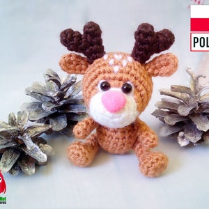 May include: A crocheted reindeer with brown antlers, a pink nose, and white accents. The reindeer is sitting in front of two silver-painted pine cones. The image includes the text "Little Owls Hut Crochet Patterns".