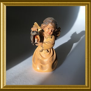 Può includere: Una piccola statuina di angelo in legno che tiene una lanterna con una candela accesa al suo interno. L'angelo indossa un vestito color crema con dettagli dorati e ha un viso delicato dipinto.