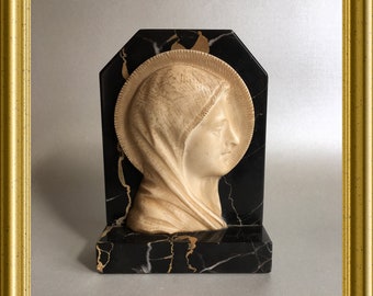 May include: A religious sculpture featuring a profile of a woman's face, carved in a cream color, set against a black marble background with gold veining. The sculpture is framed in a gold-colored frame.