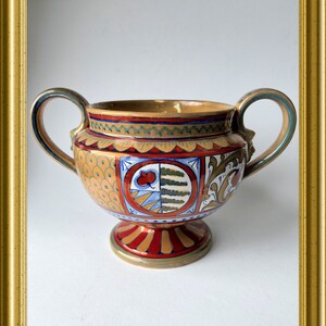 Op de afbeelding: Een decoratieve keramische urn met twee handvatten, met een gedetailleerd, kleurrijk ontwerp. De urn heeft een gouden binnen- en buitenkant met rode, blauwe en bruine accenten. De basis is rood en goud, en de handvatten zijn blauw en goud.