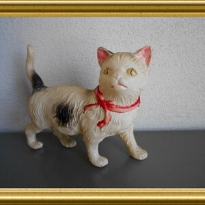 May include: A vintage cream-colored cat figurine with black spots and a red ribbon collar. The cat has yellow eyes and pink inner ears. The figurine is set against a neutral background and framed in gold.