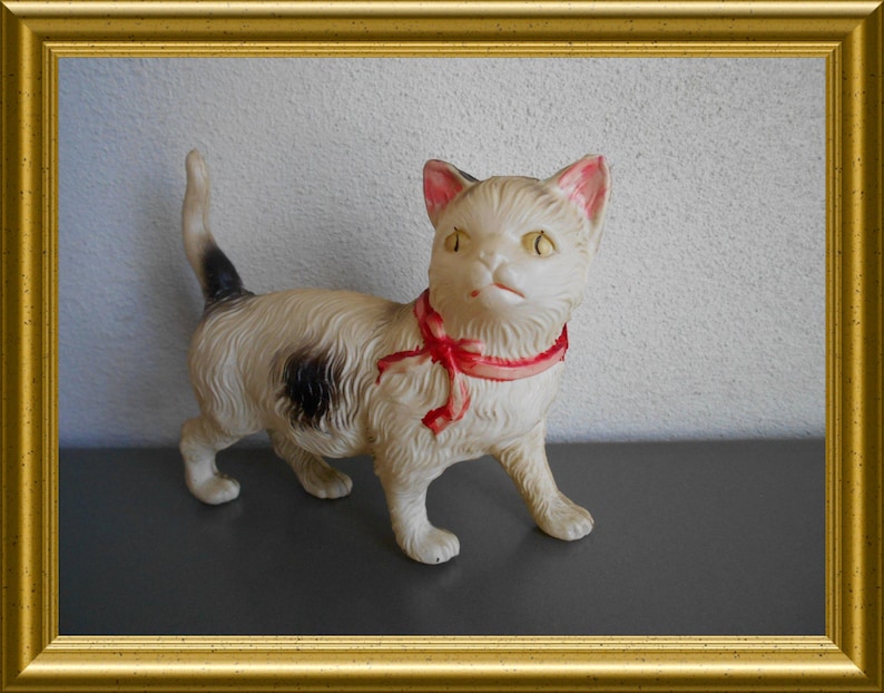 May include: A vintage cream-colored cat figurine with black spots and a red ribbon collar. The cat has yellow eyes and pink inner ears. The figurine is set against a neutral background and framed in gold.