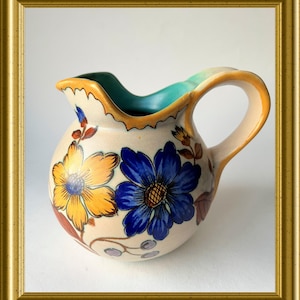Pode incluir: Jarro de cerâmica com base creme, adornado com desenhos florais amarelos e azuis pintados à mão. O jarro tem uma alça curva e uma borda ondulada com interior turquesa. O jarro é emoldurado em uma moldura dourada.