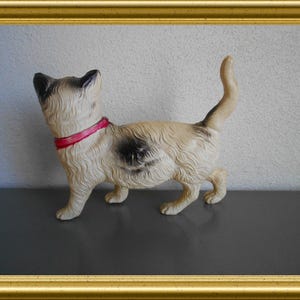 May include: A vintage cat figurine with a cream-colored body, black markings, and a red collar. The cat is standing with its tail up, set against a neutral background and framed in gold.