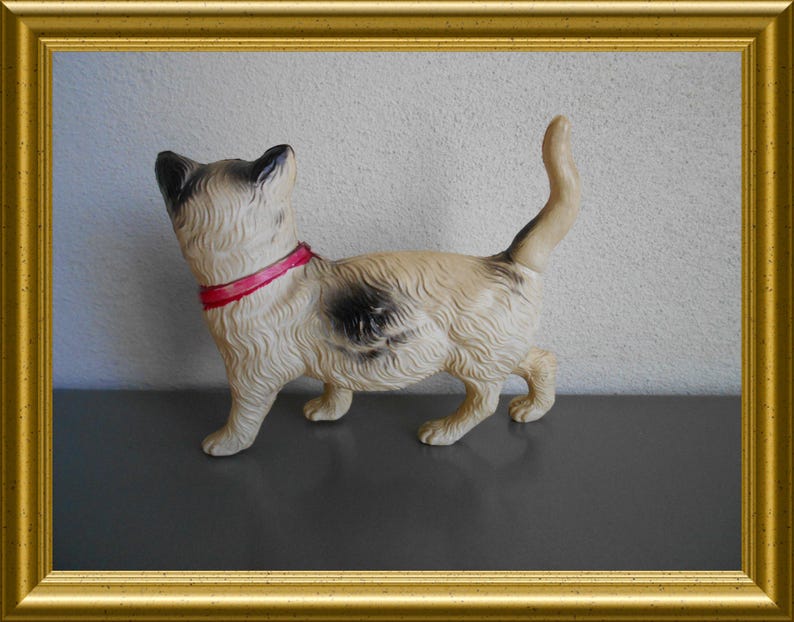 May include: A vintage cat figurine with a cream-colored body, black markings, and a red collar. The cat is standing with its tail up, set against a neutral background and framed in gold.