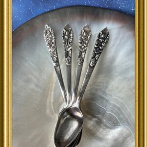 Op de afbeelding: Vier antieke zilveren lepels met sierlijke bloemmotieven op de handvatten zijn in een waaier gerangschikt. De lepels staan tegen een parelmoer achtergrond, omlijst in een goudkleurige lijst. De achtergrond is blauw.