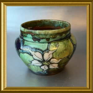 Può includere: Un vaso in ceramica fatto a mano con un'ampia apertura e un corpo arrotondato. Il vaso presenta un motivo floreale con petali bianchi e verdi, accentuato da dettagli blu e marroni. L'esterno ha una finitura lucida.