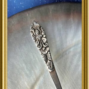 Op de afbeelding: Close-up van een zilveren keukengerei met sierlijke bloemendetails op het handvat. Het keukengerei staat tegen een parelmoer achtergrond, omlijst in goud. Het ontwerp van het handvat bevat ingewikkelde gravures van bloemen en bladeren.