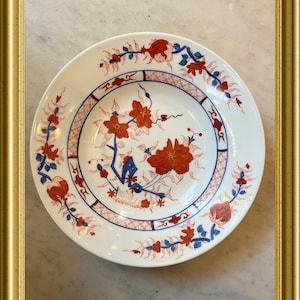 Puede incluir: Plato de cerámica blanca con un diseño floral en rojo y azul. El plato presenta un motivo floral central y un borde con patrones florales y geométricos repetidos. El plato está enmarcado en un marco dorado.