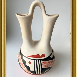 Pode incluir: Um vaso de cerâmica creme com dois bicos e uma alça curva. O vaso apresenta um design geométrico em preto, vermelho e branco ao redor da base. O vaso é exibido em uma moldura dourada.