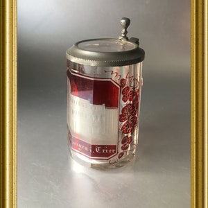 May include: A vintage glass beer stein with a red and white design. The stein features a depiction of a building and the text "Mosel. Trier".