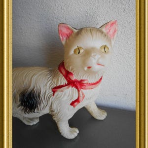May include: A vintage white cat figurine with a red ribbon around its neck. The cat has yellow eyes, pink-lined ears, and a black spot on its side. The figurine is set against a neutral background and framed in gold.