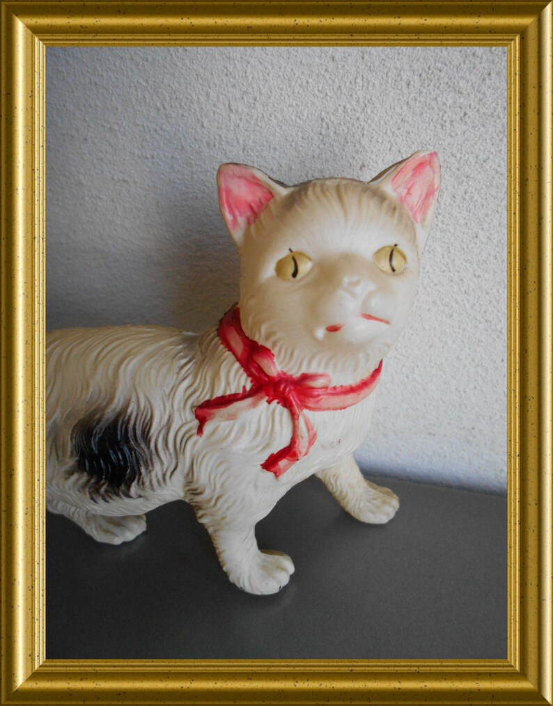 May include: A vintage white cat figurine with a red ribbon around its neck. The cat has yellow eyes, pink-lined ears, and a black spot on its side. The figurine is set against a neutral background and framed in gold.