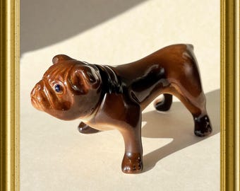 May include: A brown ceramic bulldog figurine with a glossy finish. The figurine is standing on all four legs with its head tilted to the side. The bulldog has a blue eye and a black nose.