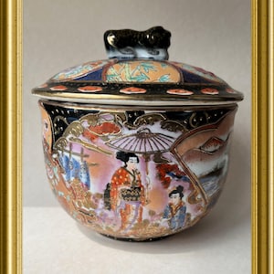 Pode incluir: Um pote de porcelana com tampa e um botão de leão preto. O pote é decorado com um design colorido de inspiração japonesa que apresenta mulheres com roupas tradicionais, flores e uma paisagem.