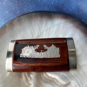May include: A rectangular wooden box with a polished dark brown finish and silver-toned metal accents on the sides. The lid features an inlaid silver-coloured metal plaque depicting a landscape with two buildings, trees, and a central figure.
