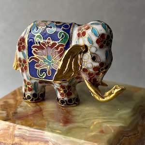 May include: A small decorative elephant figurine crafted with white, blue, and red cloisonné enamel featuring floral patterns and gold-toned metal accents on the ears and tusks. The elephant is standing on a polished, multi-colored stone base.