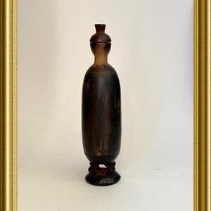 Op de afbeelding: Een donkerbruine houten fles met een klein, rond, houten deksel. De fles heeft een afgerond lichaam en een kleine, ronde basis met drie poten.