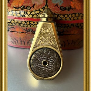 May include: An antique gold-coloured perfume bottle with detailed engraved floral and geometric designs. The bottle has a hexagonal shape with a round, patterned base and a conical top. The background features a decorative, lacquered box with red, black, and gold accents.