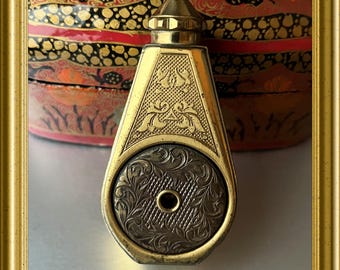 May include: A gold-toned antique perfume bottle with intricate engraved floral and geometric patterns. The bottle has a hexagonal shape with a round, patterned base and a conical top. The background features a decorative, lacquered box with red, black, and gold accents.
