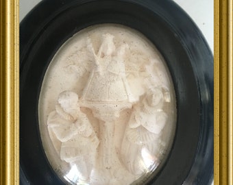 May include: A white cameo framed in a black oval frame with a gold frame around the outside. The cameo depicts a religious scene with three figures.