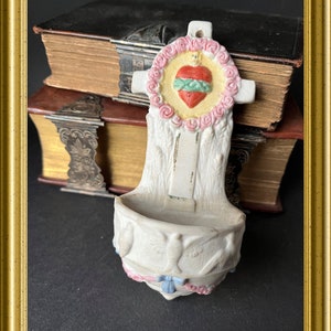 May include: A white ceramic holy water font with a cross-shaped top featuring a heart design. The font has a basin with dove and floral details, accented with pink and blue ribbons. The background includes antique books.
