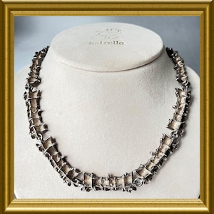 Op de afbeelding: Een zilverkleurige ketting met een ingewikkeld, getextureerd ontwerp. De ketting wordt gepresenteerd op een witte buste, omlijst in goud. De merknaam "astrelle" is zichtbaar op de buste.