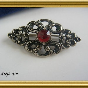 May include: A silver filigree brooch with a red gemstone in the center. The brooch is in the shape of a teardrop and has a delicate design.