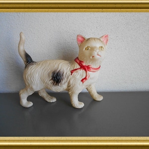 May include: A vintage cat figurine with a cream-colored body, black spot, and a red ribbon around its neck. The cat has pink-lined ears and a black and white tail. The figurine is displayed in a gold frame.