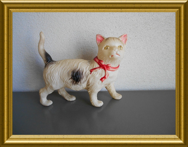 May include: A vintage cat figurine with a cream-colored body, black spot, and a red ribbon around its neck. The cat has pink-lined ears and a black and white tail. The figurine is displayed in a gold frame.