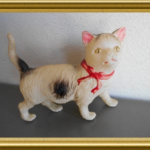 May include: A vintage cat figurine with a cream-colored body, black spot, and a red ribbon around its neck. The cat has pink ears and yellow eyes. The figurine is displayed in a gold frame.