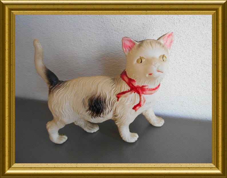 May include: A vintage cat figurine with a cream-colored body, black spot, and a red ribbon around its neck. The cat has pink ears and yellow eyes. The figurine is displayed in a gold frame.