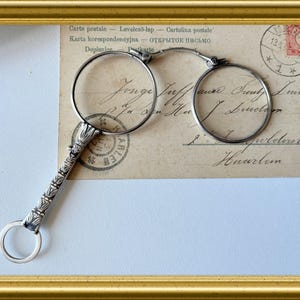 Op de afbeelding: Antieke zilverkleurige bril met een gedetailleerde handgreep en ronde lenzen, rustend op een vintage ansichtkaart. De ansichtkaart bevat handgeschreven tekst, postzegels en tekst in meerdere talen. De afbeelding is omlijst in een goudkleurige lijst.
