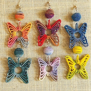 以下が含まれることがあります： 丸いビーズがついたカラフルな蝶の形をしたイヤリングが6ペア。イヤリングは編まれた紐で作られており、赤、オレンジ、黄色、緑、青、紫など、さまざまな色があります。