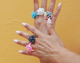 May include: A hand model wearing four beaded flower rings. The rings are in shades of blue, white, rainbow, black, and pink.