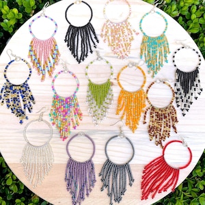 May include: A collection of thirteen circular hoop earrings featuring dangling fringe made from small glass beads in various colors including black, white, red, blue, green, orange, yellow, purple, and metallic gold, arranged on a round wooden surface.