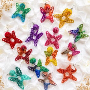 Peut inclure: Une collection de boucles d'oreilles en forme de papillon fabriquées en fil coloré. Chaque boucle présente une ouverture circulaire centrale, des motifs d'ailes en boucles complexes et une petite perle sphérique au sommet. Couleurs variées.