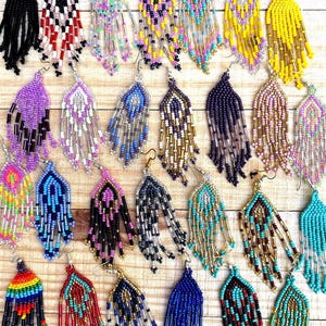 May include: A collection of beaded earrings in various colors, including black, red, blue, yellow, and turquoise. The earrings feature intricate designs with dangling strands of beads, showcasing a bohemian style. The earrings are displayed on a light wooden surface.