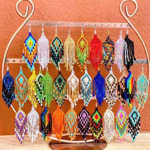 May include: A collection of thirty-two pairs of handcrafted beaded fringe earrings displayed on a copper-toned metal stand. Each pair features unique geometric patterns and vibrant color combinations, including solid, striped, and multi-colored designs.