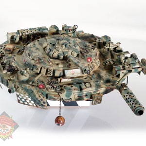 May include: A detailed model tank with a camouflage pattern in shades of green, brown, and tan. The model features a long gun barrel, multiple turrets, and intricate detailing. A small red star is visible on the side. A decorative ball hangs from the bottom.