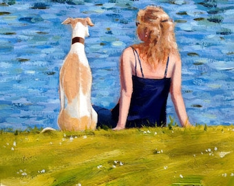 May include: A woman with long blonde hair sits on a grassy bank with a white and tan dog. The dog is looking at the water, while the woman is looking at the dog. The water is blue and rippling.