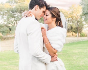 May include: A bride and groom in white wedding attire embrace and look at each other. The bride is wearing a white fur shawl and the groom is wearing a white tuxedo jacket.