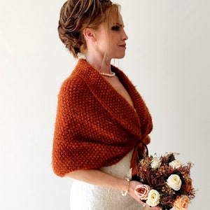 May include: A rust-colored, textured knit shawl with a wide collar and a brown ribbon tie closure. The garment features a fuzzy, looped yarn construction and is worn over a white lace dress, paired with a bouquet of cream and peach roses.