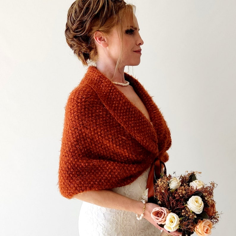 May include: A rust-colored, textured knit shawl with a wide collar and a brown ribbon tie closure. The garment features a fuzzy, looped yarn construction and is worn over a white lace dress, paired with a bouquet of cream and peach roses.