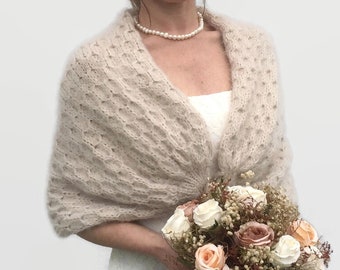 May include: A beige knitted shawl with a delicate openwork pattern. The shawl is draped over the shoulders of a person wearing a white dress and holding a bouquet of flowers.