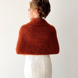 May include: A rust-colored, textured knit bolero shrug featuring a wide collar and a short, cropped silhouette. The garment has a fuzzy, nubby surface texture and covers the shoulders and upper back, worn over a white lace dress.