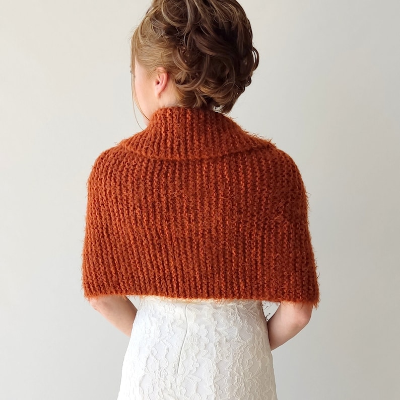 May include: A rust-colored knitted shrug featuring a textured ribbed pattern and a wide folded collar. The garment is worn over a white lace dress, covering the shoulders and upper back with a soft, fuzzy yarn construction.