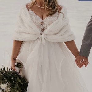 May include: A white faux fur shawl with a satin ribbon tie. The shawl is draped over a white wedding dress with lace detailing. The bride is holding a bouquet of white flowers and greenery.