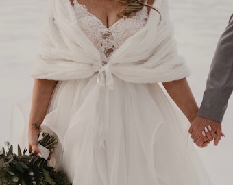 May include: A white faux fur shawl with a satin ribbon tie. The shawl is draped over a white wedding dress with lace detailing. The bride is holding a bouquet of white flowers and greenery.