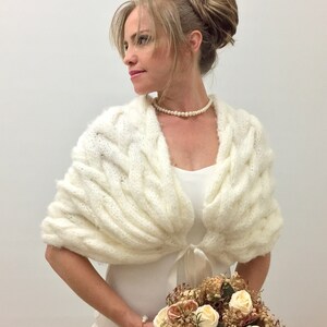 May include: White knitted bridal shawl with a cable knit pattern and a satin ribbon tie. The shawl is draped over the shoulders of a person wearing a white dress.